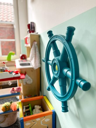 Blaues Steuerrad an der Wand einer Kita neben einer Spielküche und Spielzeugkisten