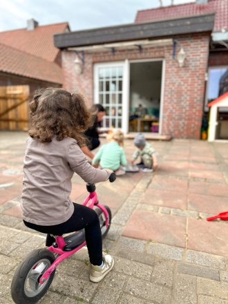 Kind auf pinkem Laufrad fährt über den Hof vor einer Kita, im Hintergrund spielen weitere Kinder