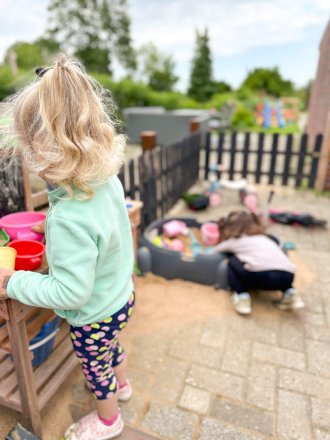Zwei Kinder spielen draußen mit Sandspielzeug im Garten, eines steht an einem Regal, das andere sitzt am Sandkasten