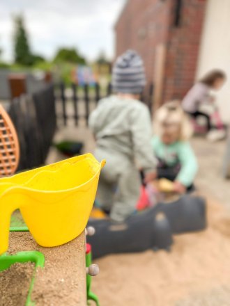 Nahaufnahme einer gelben Sandschaufel mit spielenden Kindern im Hintergrund im Sandkasten
