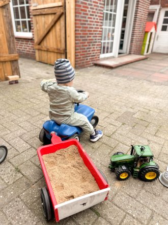 Kleinkind fährt auf einem blauen Rutschfahrzeug mit Sandanhänger im Hof